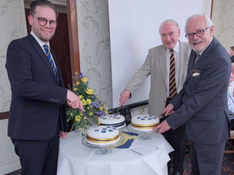 Luke Anderson, Richard Anderson and James Loader each standing holding knives and ceremonially cutting the three cakes, each of which is decorated with a digit, together making "125"